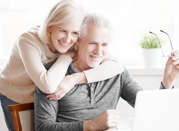 Elderly man holds glasses while wife hugs him while smiling at laptop