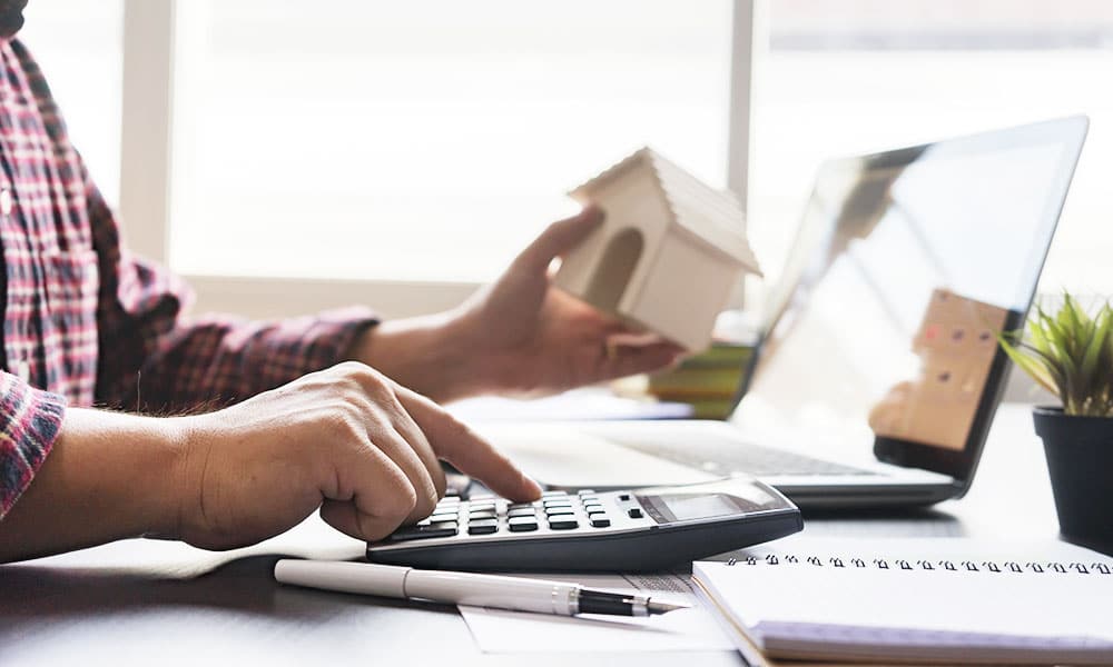 Man typing on calculator with wooden built toy house in hand